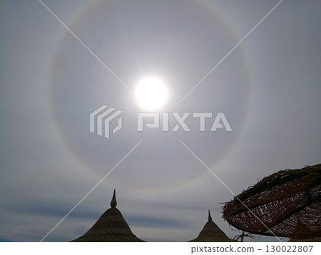 Sun with a Halo Over a Beach with Woven Umbrellas and a Cloudy Sky 130022807