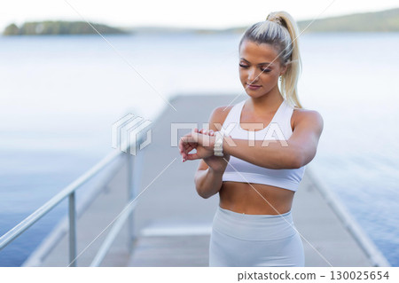Woman Monitoring Workout Progress with Smartwatch on a Lakeside Boardwalk 130025654