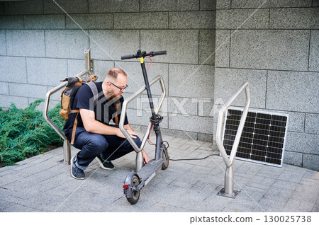 Man connects solar panel to electric scooter for charge, highlighting practical application of sustainable renewable energy. Concept of integration of eco-friendly technology in urban transportation. 130025738