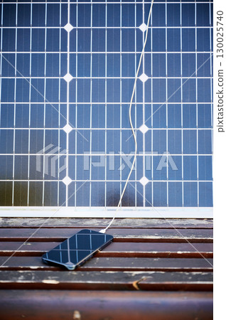 Solar panel placed on wooden bench, with cable connected to smartphone. Makeshift setup for charging phone using solar energy, highlighting portable and renewable power solutions. Solar panel placed on wooden bench, with cable connected to smartphone. Makeshift setup for charging phone using solar energy, highlighting portable and renewable power solutions. 130025740