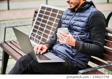 Cropped view of man sits on wooden bench in urban setting, working on laptop with cup of coffee in hand. Solar panel rests beside, emphasizing sustainable, mobile workspace. Cropped view of man sits on wooden bench in urban setting, working on laptop with cup of coffee in hand. Solar panel rests beside, emphasizing sustainable, mobile workspace. 130025746