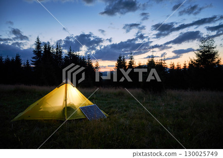 Glowing orange tent illuminated at dusk beside solar panel in grassy field. Backdrop of silhouetted trees and starry night sky emphasizes eco-friendly and serene camping experience. 130025749