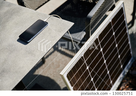 Smartphone connected to solar panel on outdoor table, emphasizing practical use of solar energy for charging devices. Integration of renewable energy, showcasing portable solar technology. 130025751