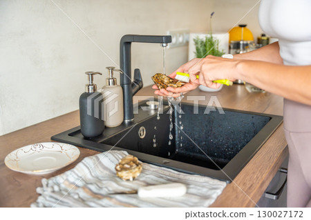 Female hands washing a fresh oyster under running tap water before opening. Hygiene and safety precautions when preparing shellfish at home. 130027172