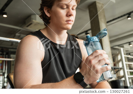 Sporty caucasian man wearing blank sleeveless tank top with water bottle in hand at gym. Relaxation after workout. Water balance. Close up. 130027261