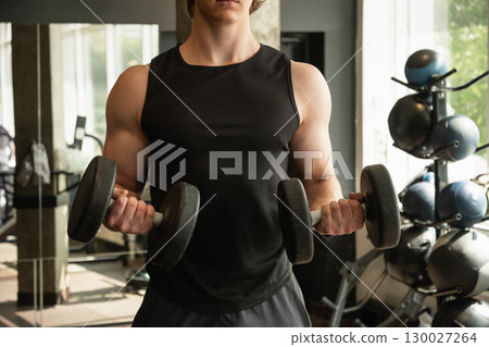 Close up of muscular arm lifting dumbbells in gym setting. Weightlifting exercises, physical fitness. Bodybuilder lifting dumbbells, biceps exercise in gym. Close up of muscular arm lifting dumbbells in gym setting. Weightlifting exercises, physical fitness. Bodybuilder lifting dumbbells, biceps exercise in gym. 130027264