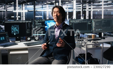 Data center admin waving hand, filming presentation tour, showcasing mainframes and AI technology. Upbeat Asian man in server room presenting artificial intelligence neural networks tech, camera A 130027290
