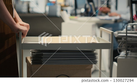 Professional archivist uses rolling trolley to transport documents and records, assisting the ceo and coworkers with efficient handling of critical paperwork in a modern workplace. Camera B. Professional archivist uses rolling trolley to transport documents and records, assisting the ceo and coworkers with efficient handling of critical paperwork in a modern workplace. Camera B. 130027294