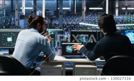 Server room system administrators using AI to review infrastructure diagnostics. IT professionals in data center analyzing performance data using artificial intelligence technology, camera A 130027316