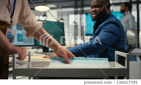 Professional archivist uses rolling trolley to transport documents and records, assisting the ceo and coworkers with efficient handling of critical paperwork in a modern workplace. 130027395