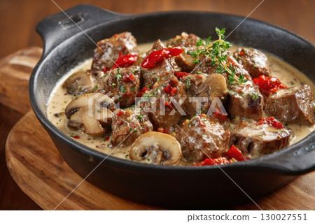 Meat and mushrooms. Dish of stir-fried beef fillet and mushrooms in a creamy thick sauce, served on an iron plate on a wooden board, close-up view. Gourmet food. Beef stroganoff large pieces. Stir Fry 130027551