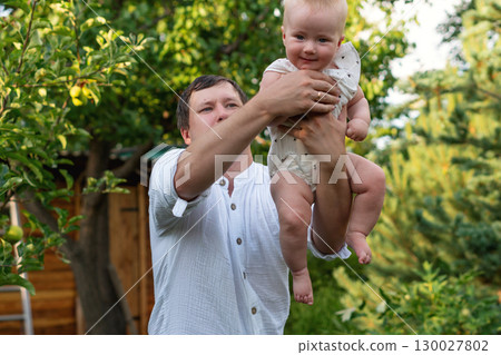Father lifts smiling baby forward amid leafy garden trees Father lifts smiling baby forward amid leafy garden trees 130027802