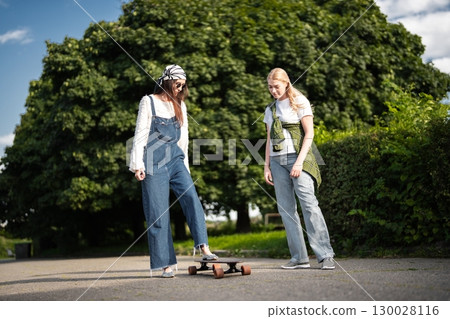 Two friends enjoying a sunny day while trying out a skateboard together Two friends enjoying a sunny day while trying out a skateboard together 130028116