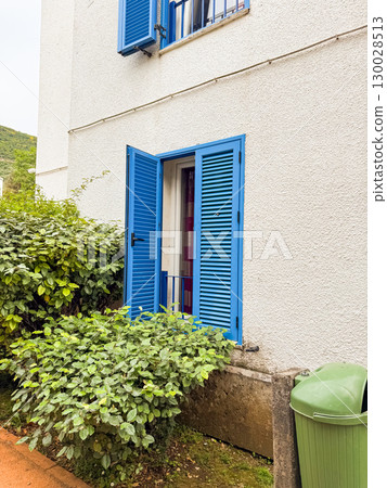 Open blue shuttered window on white building with surrounding bushes and greenery. Residential architecture, lifestyle, eco balance and summer outdoor neighborhood environment. 130028513