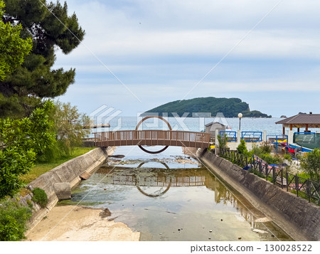 Wooden bridge with circular design reflected in water facing small island in sea. Travel, tourism, architecture, leisure and scenic coastal destination with unique design. 130028522