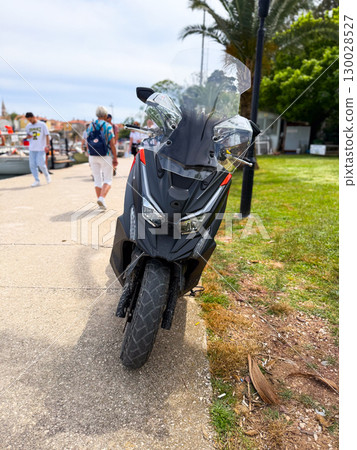 Black scooter with windshield parked on a coastal promenade near people and palm. Urban lifestyle, freedom, holiday transport and modern travel mobility. Black scooter with windshield parked on a coastal promenade near people and palm. Urban lifestyle, freedom, holiday transport and modern travel mobility. 130028527