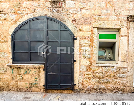 Street view of an ATM built into an old stone wall with black door nearby. Reflects urban finance, commerce and the concept of money access. 130028532