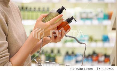 Woman selecting shampoo, body wash, or hand soap in a cosmetics store, shopping basket in hand. Female customer reading labels and comparing products. Concept of conscious choice and personal care. 130029600