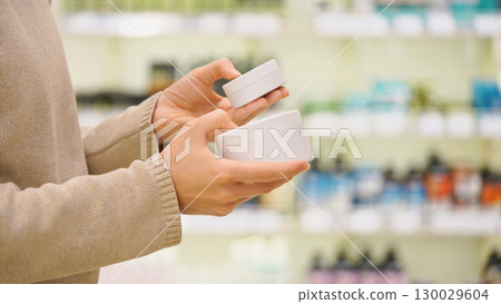 Woman selecting skincare cream for face or body or a hair mask in a beauty store, reading ingredients, comparing products. Female customer holding and examining cosmetic jar. Concept of personal care. Woman selecting skincare cream for face or body or a hair mask in a beauty store, reading ingredients, comparing products. Female customer holding and examining cosmetic jar. Concept of personal care. 130029604