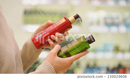 Customer comparing haircare products in a beauty store. Woman reading ingredient labels on bottles of shampoo, conditioner or balm. Concept of conscious choice and personal care for hair and scalp. Customer comparing haircare products in a beauty store. Woman reading ingredient labels on bottles of shampoo, conditioner or balm. Concept of conscious choice and personal care for hair and scalp. 130029606