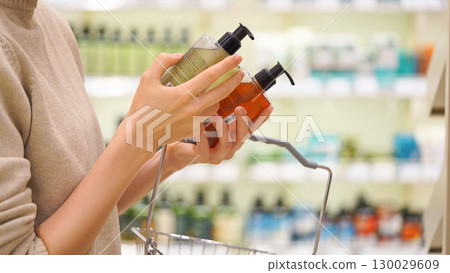 Woman selecting shampoo, body wash, or hand soap in a cosmetics store, shopping basket in hand. Female customer reading labels and comparing products. Concept of conscious choice and personal care. 130029609