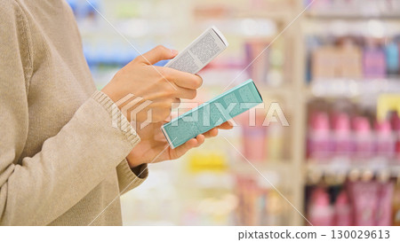 Woman selecting a cosmetic serum for face or hair in a beauty store, reading ingredients, comparing products. Female customer holding skincare or haircare item. Concept of conscious choice, self-care. 130029613
