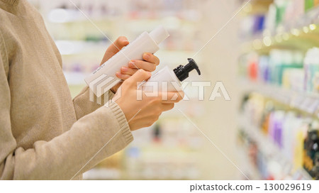 Customer choosing facial cleanser in a cosmetics store. Woman comparing gel, foam or enzyme powder for skincare. Reading ingredients. Close-up hands holding bottles. Concept of conscious beauty choice 130029619
