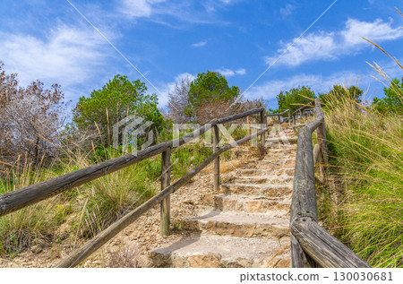 Stone staircase and wooden fence leading up to hill with green grass, bushes and trees in Serra Gelada natural park in summer day, Valencian Community, Spain 130030681