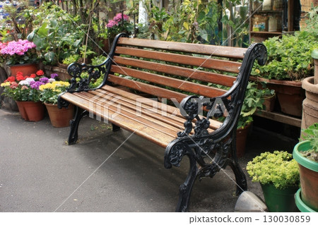 Empty brown metal and wooden bench with no people in a public park, with trees and bushes of flowers in the background, front view. Vacant bench in the backyard garden among a blooming flowers. Empty brown metal and wooden bench with no people in a public park, with trees and bushes of flowers in the background, front view. Vacant bench in the backyard garden among a blooming flowers. 130030859