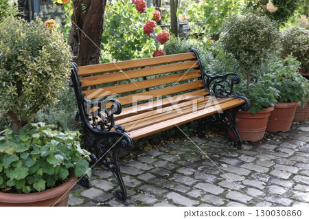 Empty brown metal and wooden bench with no people in a public park, with trees and bushes of flowers in the background, front view. Vacant bench in the backyard garden among a blooming flowers. Empty brown metal and wooden bench with no people in a public park, with trees and bushes of flowers in the background, front view. Vacant bench in the backyard garden among a blooming flowers. 130030860