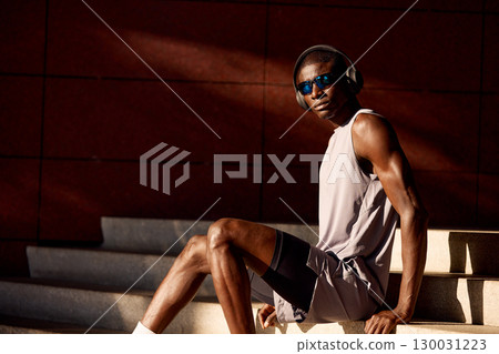 Young athletic man resting on stairs wearing headphones and sunglasses outdoors 130031223