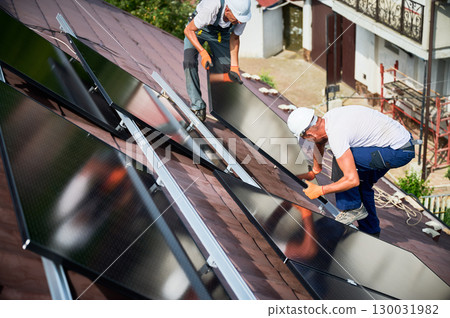 Workers building solar panel system on roof of house. Two men installers in helmets carrying photovoltaic solar module outdoors. Alternative, green and renewable energy generation concept. Workers building solar panel system on roof of house. Two men installers in helmets carrying photovoltaic solar module outdoors. Alternative, green and renewable energy generation concept. 130031982
