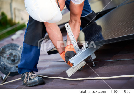 Worker building solar panel system on rooftop of house for generating electricity through photovoltaic effect. Close up man using ruler to measure mounting equipment for precise installation. Worker building solar panel system on rooftop of house for generating electricity through photovoltaic effect. Close up man using ruler to measure mounting equipment for precise installation. 130031992