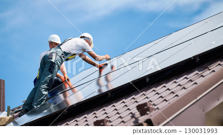 Workers building photovoltaic solar panel system on rooftop of house. Men technicians in helmets installing solar module with help of hex key outdoors. Alternative, renewable energy generation concept Workers building photovoltaic solar panel system on rooftop of house. Men technicians in helmets installing solar module with help of hex key outdoors. Alternative, renewable energy generation concept 130031993