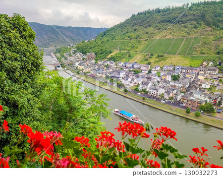 Scenic river view with colorful flowers in foreground, showcasing charming village along the water and lush green hills in the background Scenic river view with colorful flowers in foreground, showcasing charming village along the water and lush green hills in the background 130032273