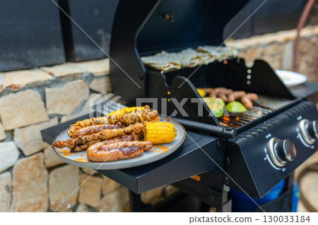 Grilled sausages, skewers, and corn on a plate near a gas barbecue with vegetables and flatbreads cooking. Outdoor summer cooking scene with delicious grilled food. 130033184