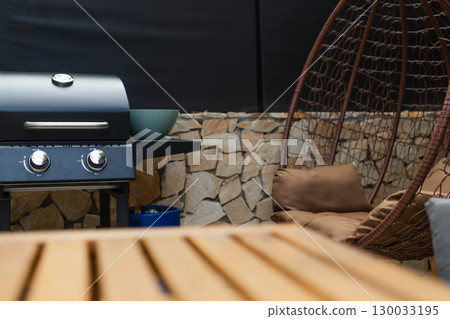 Close-up of a wooden outdoor table with a blurred gas grill in the background. Backyard BBQ setup ready for summer cooking and social gatherings. 130033195
