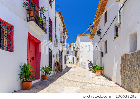 Altea old town narrow street with typical white houses buildings, flowers plants in pots in sunny summer day, Altea city historical centre Casco Antiguo, Alicante province, Valencian Community, Spain 130033292