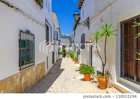 Altea old town narrow empty street with white houses traditional buildings, flowers plants in pots in sunny summer day, Altea city historical centre, Alicante province, Valencian Community, Spain 130033294
