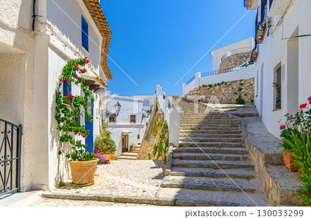 Altea old town narrow street with staircase steps, typical white houses buildings, flowers plants in pots in sunny summer day, Altea city historical centre Casco Antiguo, Alicante province, Spain 130033299