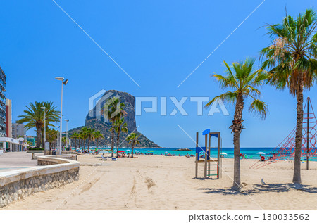 Calpe town sandy beach with palm trees on coastline Costa Blanca coast, rock mountain Penon de Ifach, Calpe bay Mediterranean Sea with azure water in sunny summer day, Alicante province, Spain 130033562