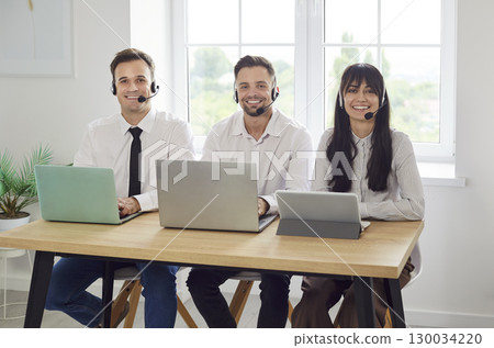 Smiling customer support agents with headsets and laptops sitting by window 130034220