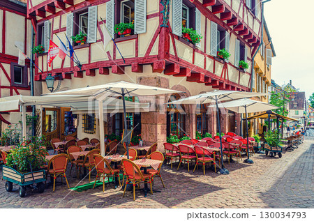 Street restaurant outdoor cafe tables, chairs and umbrellas, old colorful building half-timbered facade and shutters windows in old town Colmar city historic centre, Alsace Grand Est region, France 130034793