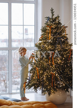 Child decorating Christmas tree with ribbons in cozy room with white wall and large window 130034951