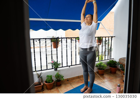 Woman Practicing Yoga on a Balcony with Plants and Blue Awning 130035030
