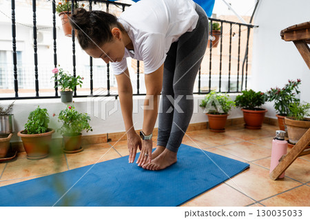 Woman Practicing Yoga Stretch on Outdoor Balcony With Green Plants and Blue Mat 130035033