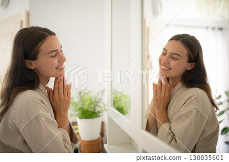 Woman smiling at herself in the mirror during morning skincare routine. Woman smiling at herself in the mirror during morning skincare routine. 130035601