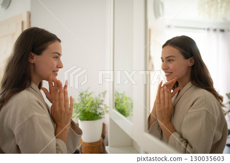 Woman smiling at herself in the mirror during morning skincare routine. 130035603