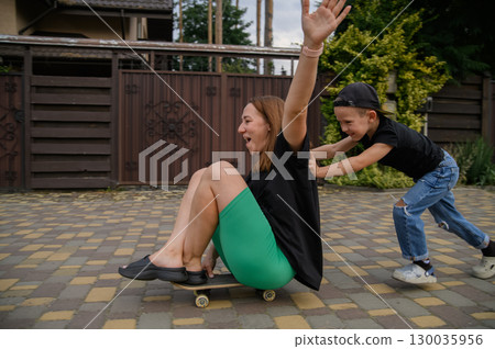 Mother Pushing Son on Skateboard in Suburban Neighborhood Street 130035956