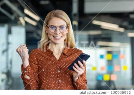 A confident woman in an office environment expresses excitement while checking her smartphone, showcasing success and positivity in business. A confident woman in an office environment expresses excitement while checking her smartphone, showcasing success and positivity in business. 130036952